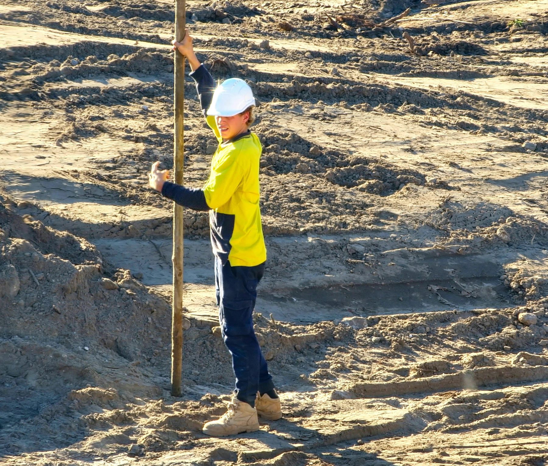 A young Australian tradie in hi-vis on a freshly graded residential site, setting up survey work in afternoon light.